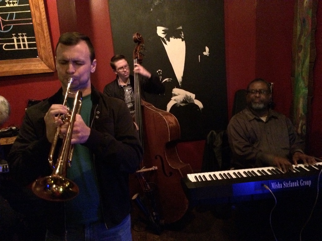 Sean McQuaid on trumpet with the Misha Stefanuk Group, an upright bassist and pianist behind him in a warm red-walled club