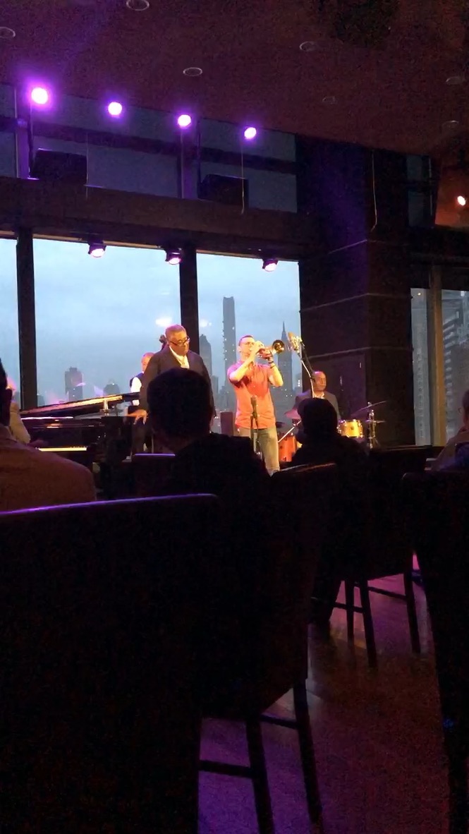 Sean McQuaid on trumpet with his quartet, the Manhattan skyline glowing behind the windows under purple stage lights